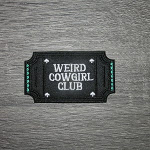 May include: A black patch with the words "WEIRD COWGIRL CLUB" in white, stylized text. The patch has a decorative border with small green accents and spade symbols. The patch is rectangular with rounded corners.