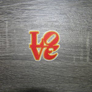 Pode incluir: Um patch bordado vermelho e amarelo com a palavra "LOVE". As letras são vermelhas com contorno amarelo e borda branca. O patch está sobre uma superfície de madeira cinza.