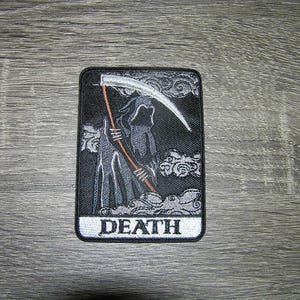 May include: Black and white embroidered patch with a skeletal figure holding a scythe. The patch has a white border and the word "DEATH" printed in white letters.