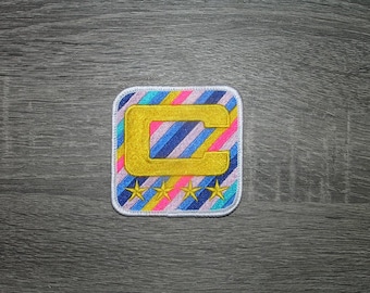 May include: A square embroidered patch with rounded corners featuring a diagonal striped background in shades of blue, pink, and yellow. A large, bold, gold-colored letter C is centered on the patch, positioned above four small gold stars.