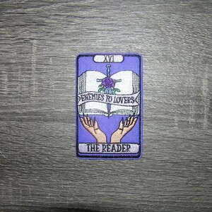 May include: Embroidered patch featuring a tarot card design. The patch is rectangular with a purple background and a white open book, a sword, and a rose. The text "ENEMIES TO LOVERS" and "THE READER" are also included.