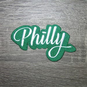 May include: A green and white embroidered patch with the word "Philly" in a cursive font. The patch has a green background with a white border and lettering. The patch is on a gray wooden surface.