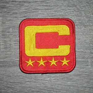 Op de afbeelding: Geborduurde patch met een rood vierkant, een geel 'C'-ontwerp en vier gele sterren eronder. Deze patch is waarschijnlijk voor kleding of accessoires.