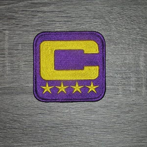 Op de afbeelding: Paarse geborduurde patch met een gouden "C" en vier gouden sterren. De patch heeft een vierkante vorm met afgeronde hoeken. Het ontwerp staat op een paarse achtergrond met textuur.