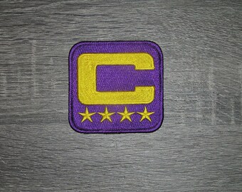 May include: Purple embroidered patch with a gold "C" and four gold stars. The patch has a square shape with rounded corners. The design is on a textured purple background.