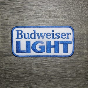 May include: Blue and white embroidered patch with the text "Budweiser Light" on a white background.