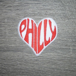 May include: A heart-shaped patch with the word "PHILLY" embroidered in red capital letters. The patch has a white background and a white border, set against a gray wooden surface.