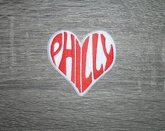 May include: A heart-shaped patch with the word "PHILLY" embroidered in red capital letters. The patch has a white background and a white border, set against a gray wooden surface.