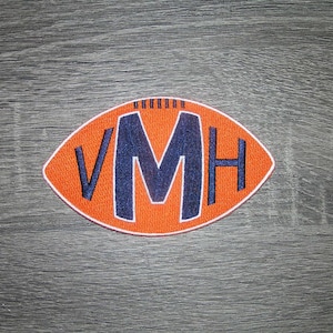 May include: Embroidered patch in the shape of a football, featuring the letters "VMH" in navy blue. The football is orange with a white border, set against a gray wood-grain background. Ideal for sports fans.