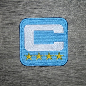 Könnte beinhalten: Ein quadratischer, gestickter Aufnäher mit hellblauem Hintergrund und einem weißen „C“ in der Mitte. Unter dem „C“ befinden sich vier goldene Sterne. Der Aufnäher hat einen hellblauen Rand.