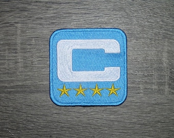 May include: A square, embroidered patch with a light blue background and a white "C" in the center. Below the "C" are four gold stars. The patch has a light blue border.