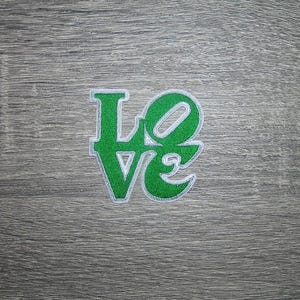 May include: A green and white embroidered patch with the word "LOVE". The letters are in a stylized font with a white border. The patch is on a gray wooden surface.