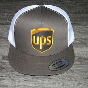 May include: A brown and white trucker hat featuring the UPS logo in gold and brown. The hat has a curved brim and a mesh back. The UPS logo is centered on the front of the hat.