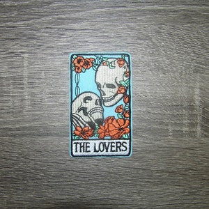 May include: An embroidered patch featuring a tarot card design. The card is blue with a white border and depicts two skeletons embracing. The card is labeled "THE LOVERS" in white text.