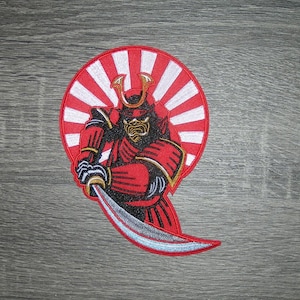 May include: Embroidered patch featuring a red samurai warrior in full armor, holding a katana. The samurai is set against a red and white sunburst design. The patch has gold and black accents.
