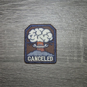 May include: Embroidered patch featuring a stylized mushroom cloud explosion design. The patch has a brown border with a blue outline. The word "CANCELED" is displayed in white block letters below the explosion.