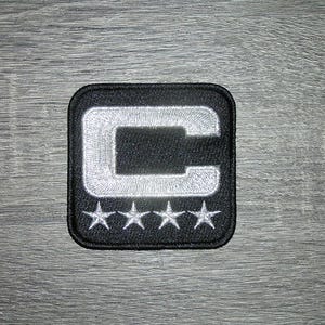 May include: A square-shaped embroidered patch featuring a black background with a large, bold white letter C centered at the top. Below the letter C, there are four small, evenly spaced white embroidered stars arranged in a horizontal row.