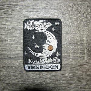 May include: Black and white embroidered patch with a crescent moon with a face and stars. The text "The Moon" is at the bottom of the patch.