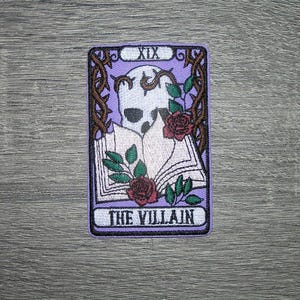 May include: A rectangular patch featuring a tarot card design. The card has a purple background with a skull, roses, and the text "THE VILLAIN". The card is framed with a brown border and thorny vines.