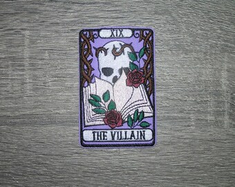 May include: A rectangular patch featuring a tarot card design. The card has a purple background with a skull, roses, and the text "THE VILLAIN". The card is framed with a brown border and thorny vines.