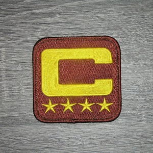 Op de afbeelding: Geborduurde patch met een gele 'C' in een kastanjebruin vierkant, met vier gele sterren eronder. De patch heeft een kastanjebruine rand, geschikt voor kleding of accessoires.