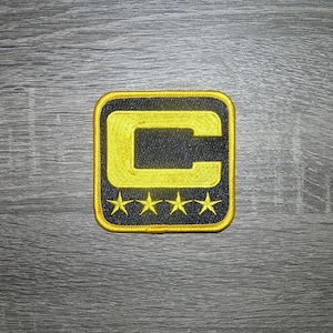 Op de afbeelding: Geborduurde patch met een gouden "C" ontwerp op een zwarte achtergrond, omlijnd in goud. Vier gouden sterren staan onder de "C". De patch heeft een vierkante vorm met afgeronde hoeken. Voor kleding of accessoires.
