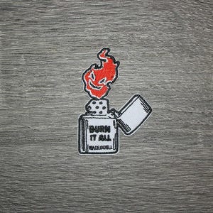 May include: Embroidered patch featuring a stylized lighter with a red flame and the text "BURN IT ALL". The patch has a white border and is set against a gray wood grain background. The text "MADE IN HELL" is also included.