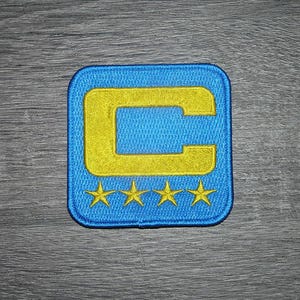 Op de afbeelding: Geborduurde patch met een blauwe vierkant, een gele "C" en vier gele sterren. De patch heeft een blauwe rand en is waarschijnlijk voor kleding of accessoires.
