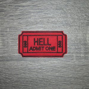 May include: A red patch with black embroidered text that reads "HELL ADMIT ONE". The patch is in the shape of a ticket with the number "666" on each side. The patch is on a gray wooden surface.