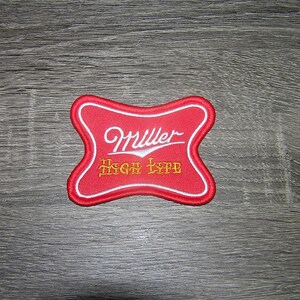 May include: A red and white embroidered patch with the Miller High Life logo. The patch has a unique shape with a white border and features the Miller logo in a cursive font and the words "High Life" in a gold, stylized font.