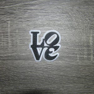 May include: A black and white embroidered patch with the word "LOVE". The letters are black with a glittery texture, outlined in white. The patch is centered on a gray wooden surface.