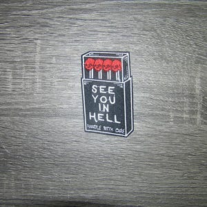 Può includere: Una toppa in bianco e nero con un design a scatola di fiammiferi. Le teste dei fiammiferi sono teschi rossi. Il testo sulla scatola di fiammiferi recita "SEE YOU IN HELL" e "HANDLE WITH CARE".