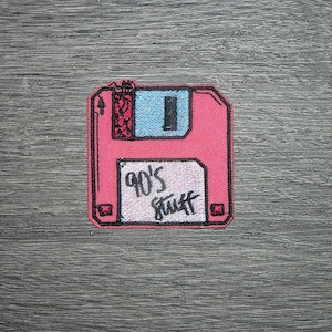 Może przedstawiać: Różowa, haftowana naszywka w kształcie dyskietki. Naszywka ma niebieski kwadrat i czarny prostokąt na górze. Tekst "90's stuff" jest napisany czarnym pismem na białym prostokącie.