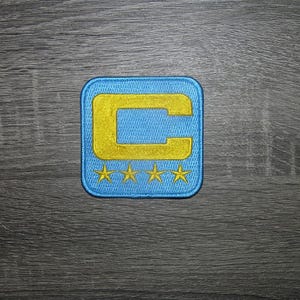 Op de afbeelding: Geborduurde patch met een lichtblauwe vierkant en een geel "C" ontwerp. Onder de "C" staan drie gele sterren. De patch is waarschijnlijk voor kleding of accessoires.