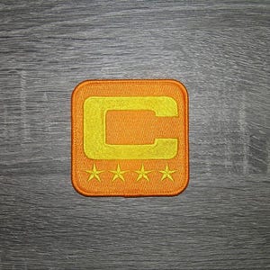May include: Embroidered patch in orange and yellow, square with rounded corners. It features a yellow letter "C" within an orange border, and four yellow stars at the bottom. Ideal for clothing.