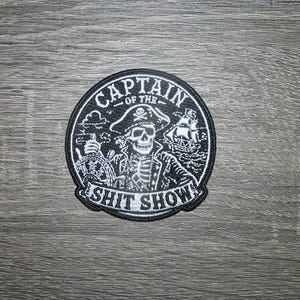 May include: A circular embroidered patch featuring a black and white design of a skeleton wearing a pirate hat and coat, holding a ship's wheel. The text CAPTAIN OF THE SHIT SHOW is embroidered in white lettering along the top and bottom borders.