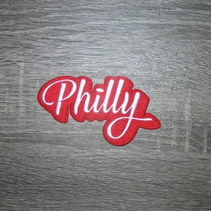 May include: An embroidered patch in red and white, featuring the word "Philly" in a cursive script. The patch has a red base with white stitching outlining the letters and border. It rests on a grey wooden surface.