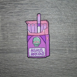 May include: A purple embroidered patch in the shape of a cigarette pack. The pack features a skull with a green mustache and the words "ALWAYS ANXIOUS" in white. The cigarettes are pink.