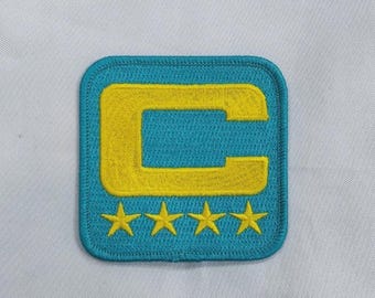 May include: A square embroidered patch with rounded corners featuring a bright yellow letter C centered on a teal background. Below the letter C, there are four small, evenly spaced yellow stars embroidered in a horizontal row on the teal fabric.