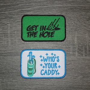 May include: Two embroidered iron-on patches. The first patch is green with black text that says "Get in the Hole" and a black golf ball with a black line through it. The second patch is white with blue text that says "Who's Your Caddy" and a blue golf bag with golf clubs.