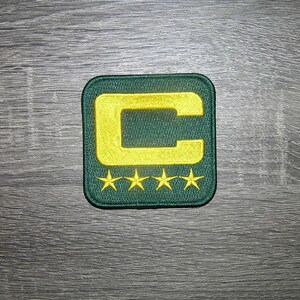 Op de afbeelding: Groen en goud geborduurde patch met een vierkante vorm. De patch heeft een grote gouden letter "C" en vier gouden sterren. De patch ligt op een grijze houten ondergrond.