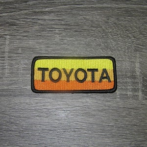 May include: A yellow and orange embroidered patch with black stitching that reads "TOYOTA".