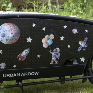 Könnte beinhalten: Ein schwarzes Lastenfahrrad mit einem Weltraum-Design. Der Laderaum ist mit Aufklebern eines Mondes, Sternen, einer Rakete, eines Astronauten und eines Raumschiffs verziert. Das Fahrrad trägt den Schriftzug "URBAN ARROW" an der Seite.