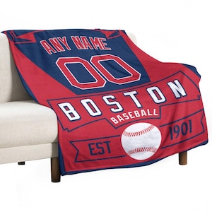Op de afbeelding: Rood en marineblauw baseballdeken met de tekst "ANY NAME" en "BOSTON BASEBALL" in het wit. Voorzien van een honkbal en het nummer "00". De deken is over een beige bank gedrapeerd.
