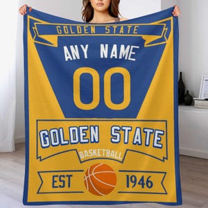 Puede incluir: Una manta de forro polar amarilla y azul con las palabras "Golden State Basketball" y "Est 1946". La manta también tiene un gráfico de baloncesto y las palabras "Any Name 00".