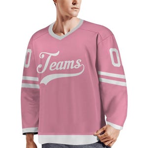 May include: Pink hockey jersey with white accents. The jersey features the word "Teams" in a cursive font across the chest, along with the number "0" on the sleeves. The jersey has a V-neck collar and white stripes on the sleeves.