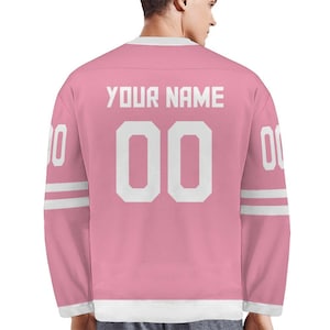 May include: Pink hockey jersey with white accents and the text "YOUR NAME" above the number "00". The jersey has white stripes on the sleeves and the number "0" on each sleeve. The jersey is made of a soft material.