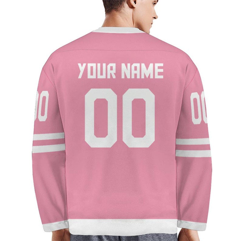 May include: Pink hockey jersey with white accents and the text "YOUR NAME" above the number "00". The jersey has white stripes on the sleeves and the number "0" on each sleeve. The jersey is made of a soft material.