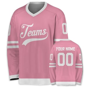 May include: Pink hockey jersey with white accents, a V-neck, and long sleeves. The front features the word "Teams" in a script font. The back displays "YOUR NAME" above the number "00".