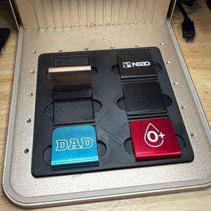 May include: An open, metallic-colored device with a black tray holding several small, rectangular objects. The objects are in various colors, including blue with the word "DAD", and red with a blood type symbol. The device has a fan and measurement markings.